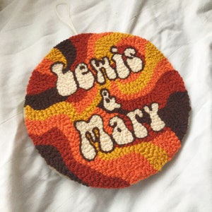 May include: A circular punch needle wall hanging with a brown, orange, and yellow swirl pattern. The text 'Lewis & Mary' is embroidered in white on the front of the wall hanging.
