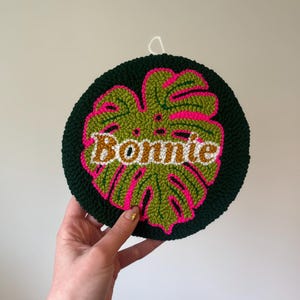 Puede incluir: Un tapiz circular de pared con un dise&ntilde;o de hoja de monstera en verde, rosa y blanco, con el nombre "Bonnie" en una fuente retro.
