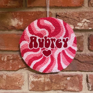 Puede incluir: Un dise&ntilde;o de espiral rosa y blanco con el nombre Aubrey en letras rojas y un peque&ntilde;o coraz&oacute;n rosa. El dise&ntilde;o est&aacute; en un lienzo redondo blanco.