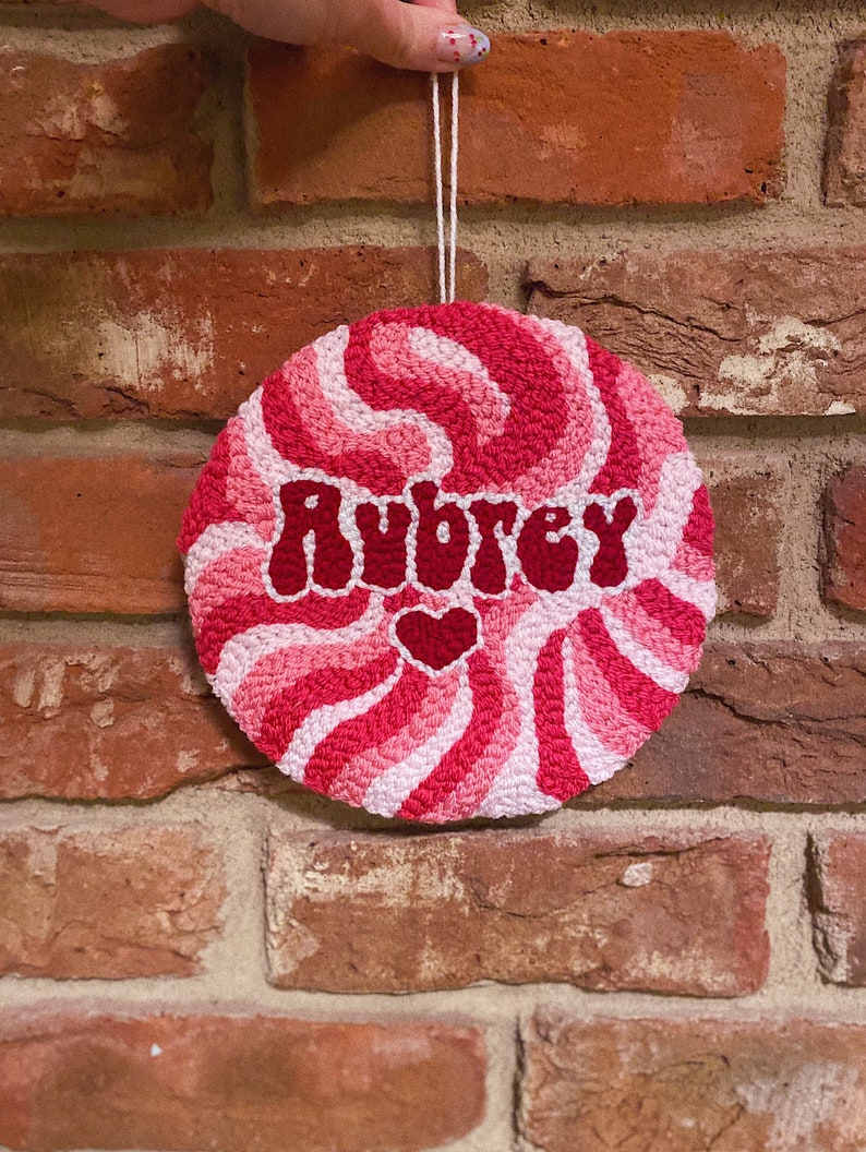 Puede incluir: Un dise&ntilde;o de espiral rosa y blanco con el nombre Aubrey en letras rojas y un peque&ntilde;o coraz&oacute;n rosa. El dise&ntilde;o est&aacute; en un lienzo redondo blanco.