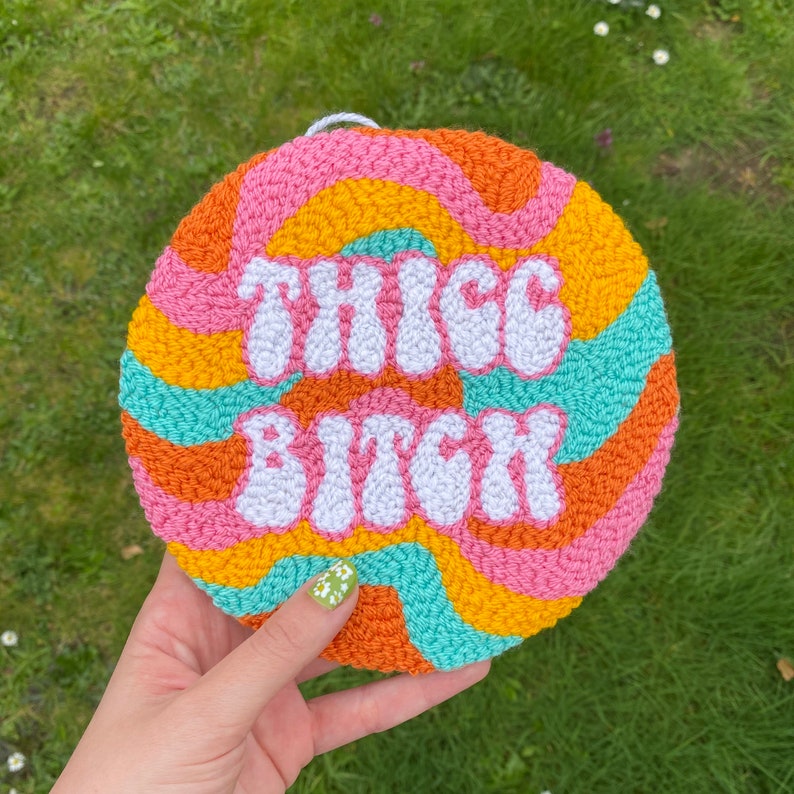 Puede incluir: Un tapiz de pared redondo, colorido y hecho de hilo con el texto "THICC BITCH" en hilo blanco.