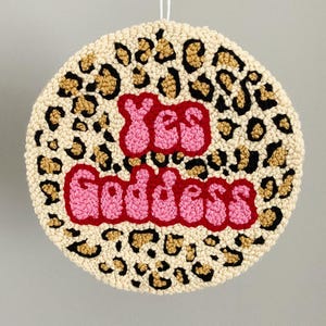 Puede incluir: Una alfombra redonda de color crema con un estampado de leopardo en negro y dorado. Las palabras "Yes Goddess" est&aacute;n bordadas en letras rosas y rojas.