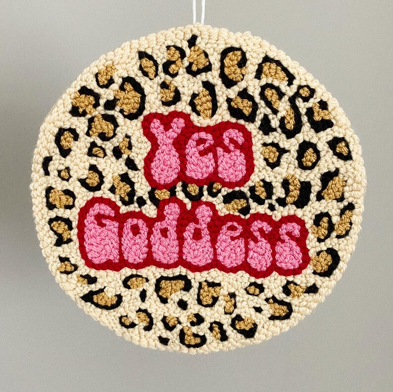 Puede incluir: Una alfombra redonda de color crema con un estampado de leopardo en negro y dorado. Las palabras "Yes Goddess" est&aacute;n bordadas en letras rosas y rojas.