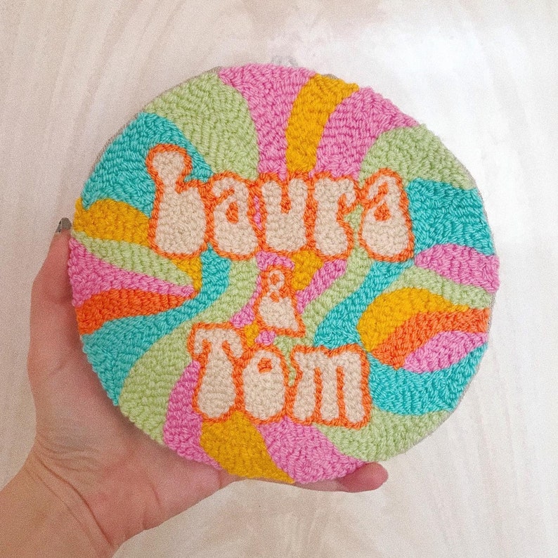 Puede incluir: Un tapiz de pared redondo, colorido y envuelto en hilo con el texto "Laura & Tom" en una fuente retro.