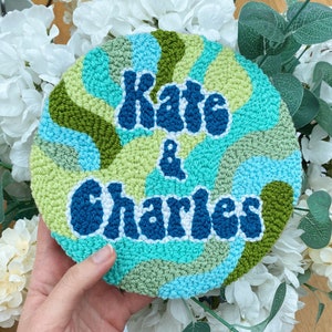 Puede incluir: Un aro de punto de aguja azul, verde y blanco con el texto "Kate & Charles" en una fuente retro.