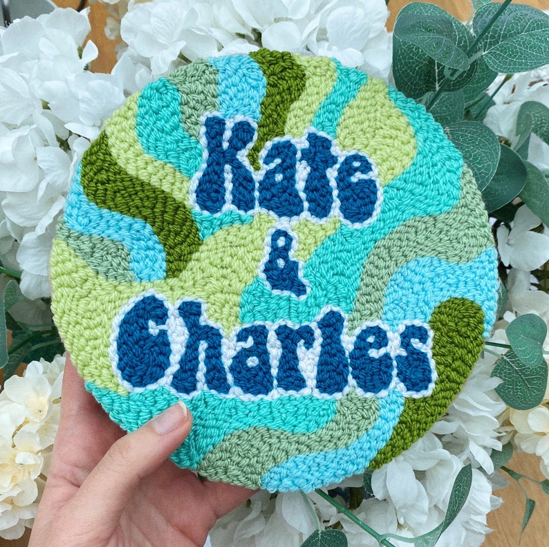 Puede incluir: Un aro de punto de aguja azul, verde y blanco con el texto "Kate & Charles" en una fuente retro.