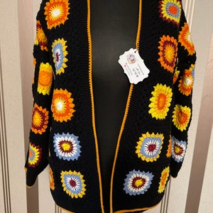 Könnte beinhalten: Schwarze Häkeljacke mit gelbem Saum und bunten Blumenmustern. Die Jacke ist vorne offen und hat lange Ärmel.