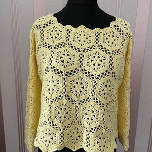 以下が含まれることがあります： フリルネックと長袖のイエローのクロシェトップ。繊細なステッチパターンでできており、花柄のデザインが施されています。