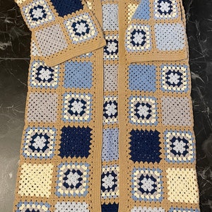 Könnte beinhalten: Ein langer, gehäkelter Cardigan mit einem Granny-Square-Muster in Beige-, Blau- und Weißtönen.