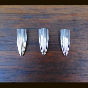May include: Three silver-colored, elongated decorative objects with a ridged design, arranged on a dark brown wooden surface. The objects have a pointed end and a wider, rounded top. The lighting creates highlights and shadows.
