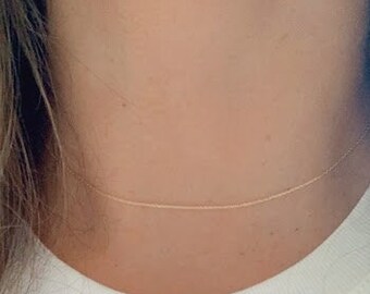 Può includere: Una semplice collana a catena d'oro indossata sul collo di una persona. La collana è sottile e delicata e viene indossata su una maglietta bianca.