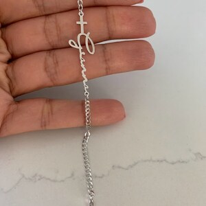Puede incluir: Un brazalete de cadena de plata con un dije de cruz y un dije con un nombre en escritura cursiva. El dije con el nombre es para el nombre "Olivia".