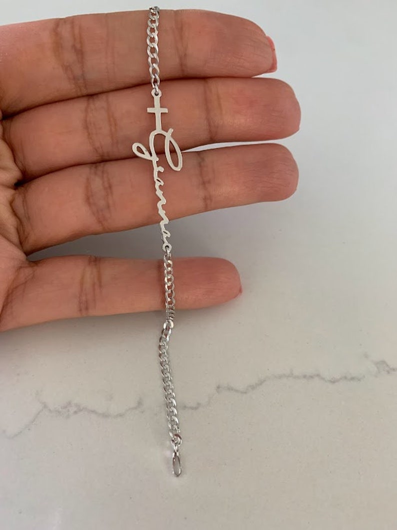 Puede incluir: Un brazalete de cadena de plata con un dije de cruz y un dije con un nombre en escritura cursiva. El dije con el nombre es para el nombre "Olivia".