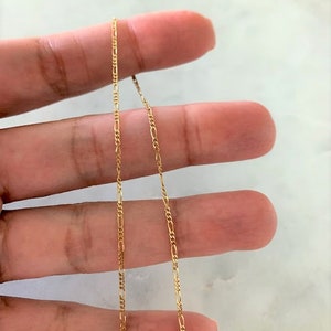 May include: A gold chain necklace with a delicate, figaro chain design. The chain is shown draped over a hand.