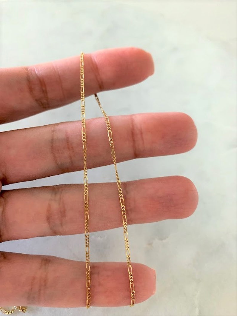May include: A gold chain necklace with a delicate, figaro chain design. The chain is shown draped over a hand.