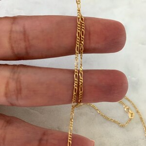 May include: A gold chain necklace with a delicate, figaro chain design. The chain is shown draped over a hand.