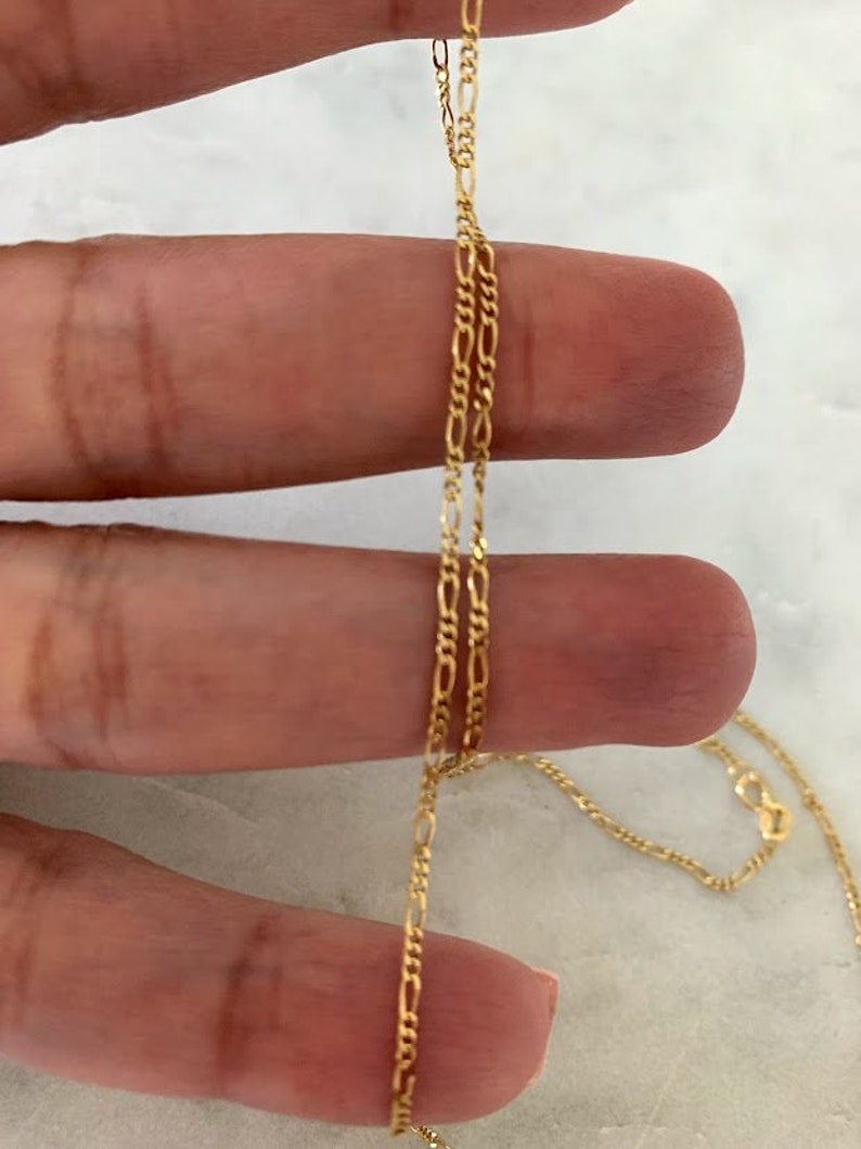 May include: A gold chain necklace with a delicate, figaro chain design. The chain is shown draped over a hand.