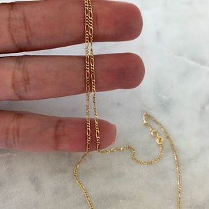May include: A gold chain necklace with a delicate, figaro chain design. The chain is shown lying on a hand and a marble surface.