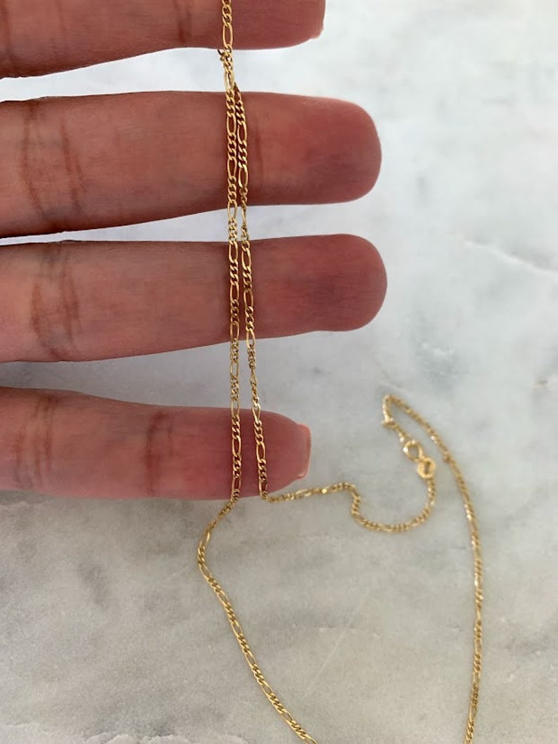 May include: A gold chain necklace with a delicate, figaro chain design. The chain is shown lying on a hand and a marble surface.