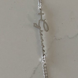 Puede incluir: Un collar de cadena de plata con un colgante que presenta una cruz y la palabra "Emma" escrita en cursiva.