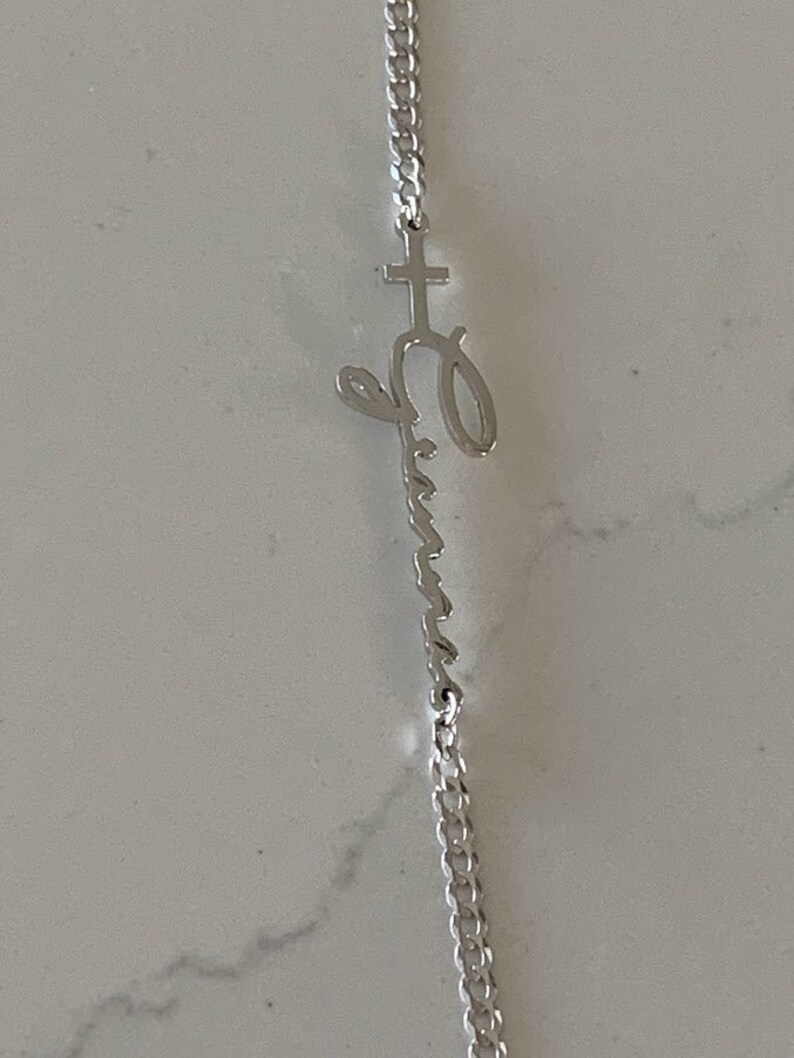 Puede incluir: Un collar de cadena de plata con un colgante que presenta una cruz y la palabra "Emma" escrita en cursiva.