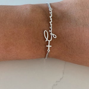 Puede incluir: Un brazalete de cadena de plata con un dije de nombre personalizado que dice "Emma" en escritura cursiva. El dije est&aacute; unido a una peque&ntilde;a cruz.