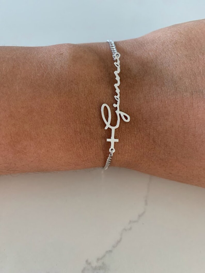 Puede incluir: Un brazalete de cadena de plata con un dije de nombre personalizado que dice "Emma" en escritura cursiva. El dije est&aacute; unido a una peque&ntilde;a cruz.