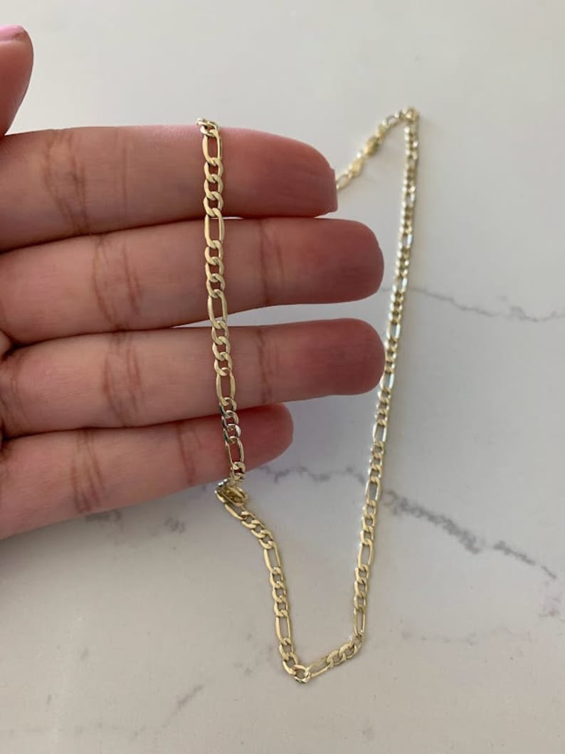 May include: A gold-colored Figaro chain necklace. The necklace features a pattern of elongated links alternating with two or three smaller, round links. The chain is displayed against a white marble surface.