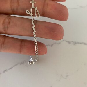 Puede incluir: Un brazalete de cadena de plata con un dije de cruz y un dije con el nombre "Abuela" escrito en cursiva.