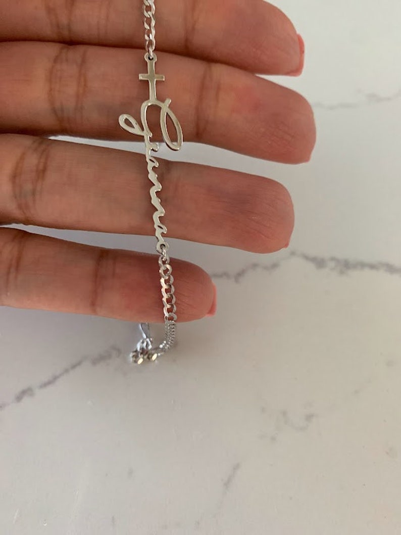 Puede incluir: Un brazalete de cadena de plata con un dije de cruz y un dije con el nombre "Abuela" escrito en cursiva.