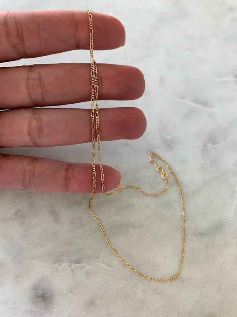 May include: A gold chain necklace with a delicate, figaro chain design. The chain is approximately 18 inches long.