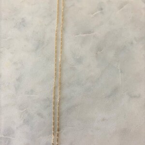 May include: A gold chain necklace with a delicate, fine link design. The chain is laying flat on a white marble surface.