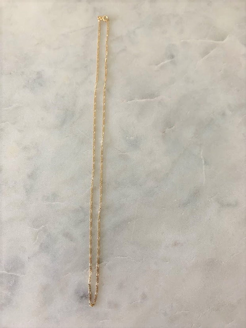 May include: A gold chain necklace with a delicate, fine link design. The chain is laying flat on a white marble surface.