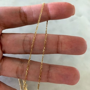 May include: A gold chain necklace with a delicate, figaro chain design. The chain is shown lying on a hand, with the ends of the chain visible.