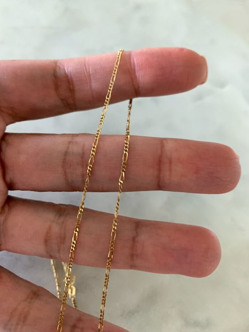 May include: A gold chain necklace with a delicate, figaro chain design. The chain is shown lying on a hand, with the ends of the chain visible.