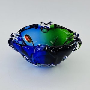 May include: A vibrant glass ashtray in shades of blue and green, with a splash of red. The ashtray has a distinctive shape with small, rounded edges. This decorative item is suitable for home decor and adds a touch of colour.