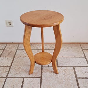 May include: A small, round wooden plant stand with a light brown finish. The stand has a circular top and a smaller circular shelf at the bottom, connected by curved legs. The stand is approximately 61 cm tall.