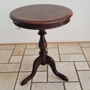 May include: A round, dark brown wooden pedestal table featuring a smooth, circular tabletop with a beveled edge. The table is supported by a central turned column that transitions into three curved, splayed legs providing a stable base.