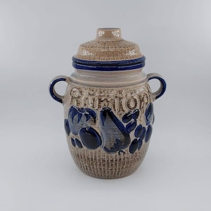 Puede incluir: Un tarro de cerámica con un diseño azul y marrón. El tarro tiene una tapa y dos asas. El tarro está decorado con un patrón de uvas y hojas.