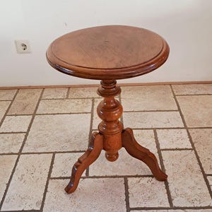 Puede incluir: Una mesa auxiliar redonda de madera con un acabado marrón pulido, que presenta una superficie circular lisa y una base de pedestal central sostenida por tres patas curvas con detalles torneados decorativos en la columna.