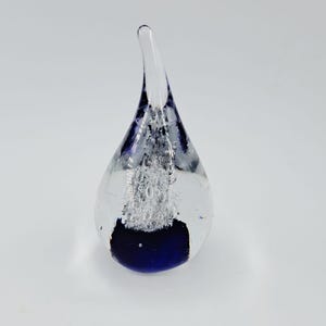 Könnte beinhalten: Ein tropfenförmiger Briefbeschwerer aus Glas mit einer klaren Außenseite und einer spitzen Oberseite. Im Inneren befindet sich eine Ansammlung kleiner Luftblasen, die über einem massiven, tiefblauen Sockel schweben und einen Kontrast bilden.