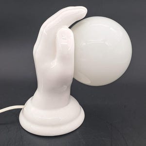 Op de afbeelding: Een witte keramische tafellamp met een sculpturale hand als voet die een bolvormige witte glazen kap ondersteunt. De lamp is voorzien van een wit elektrisch snoer dat uit de voet komt en is ontworpen om licht door de glazen bol te verspreiden.