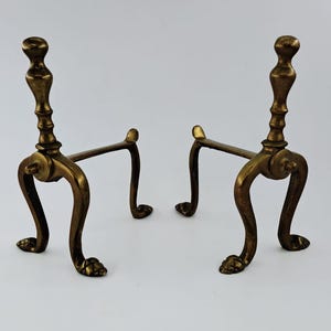 Pode incluir: Um par de tições de latão com postes verticais, remates arredondados, pernas curvas e pés em forma de garra. Estes acessórios de lareira têm um acabamento metálico dourado e uma estrutura com barras horizontais de ligação frontal.