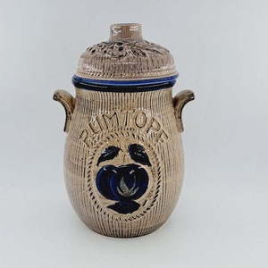 May include: A ceramic jar with a lid, decorated with a blue and white apple design and the word "RUMTORE" in blue lettering. The jar has a textured surface and is a light brown colour.