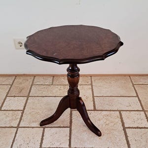 May include: A round, dark brown wooden pedestal table featuring a scalloped edge tabletop with a burl wood grain pattern. The table is supported by a central turned column post that connects to three curved legs extending outward at the base.