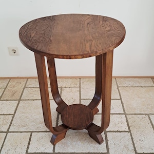 May include: A round wooden side table with a medium brown wood grain finish. The table features a circular top supported by three curved vertical legs that connect to a smaller, circular base platform centred near the floor.