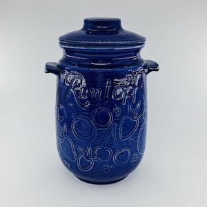 May include: A cobalt blue ceramic Rumtopf pot with a matching lid. The pot features embossed fruit designs, including cherries, strawberries, and plums. The word "Rumtopf" is embossed on the front. The pot has two side handles.