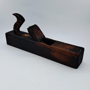 Op de afbeelding: Een vintage houten handschaaf met een donkerbruine, verweerde afwerking. Het gereedschap heeft een rechthoekig lichaam, een gebogen achterhandgreep en een centraal mesmechanisme dat is vastgezet met een houten wig, met zichtbare gebruikssporen.