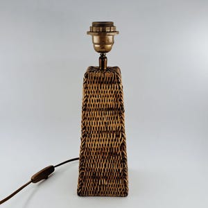 May include: A rectangular table lamp with a woven rattan base and a brass-colored bulb socket. The lamp has a brown electrical cord with an on/off switch. The lamp is a neutral color and would fit in many home decor styles.