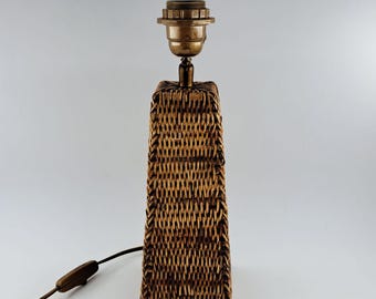 May include: A rectangular table lamp with a woven rattan base and a brass-colored bulb socket. The lamp has a brown electrical cord with an on/off switch. The lamp is a neutral color and would fit in many home decor styles.